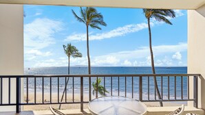 A balcony with a round table and four chairs, overlooking a beach with palm trees and the ocean.
