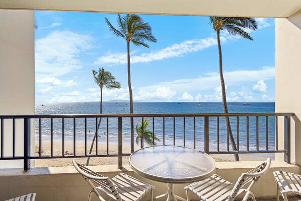 A balcony with a round table and four chairs, overlooking a beach with palm trees and the ocean.