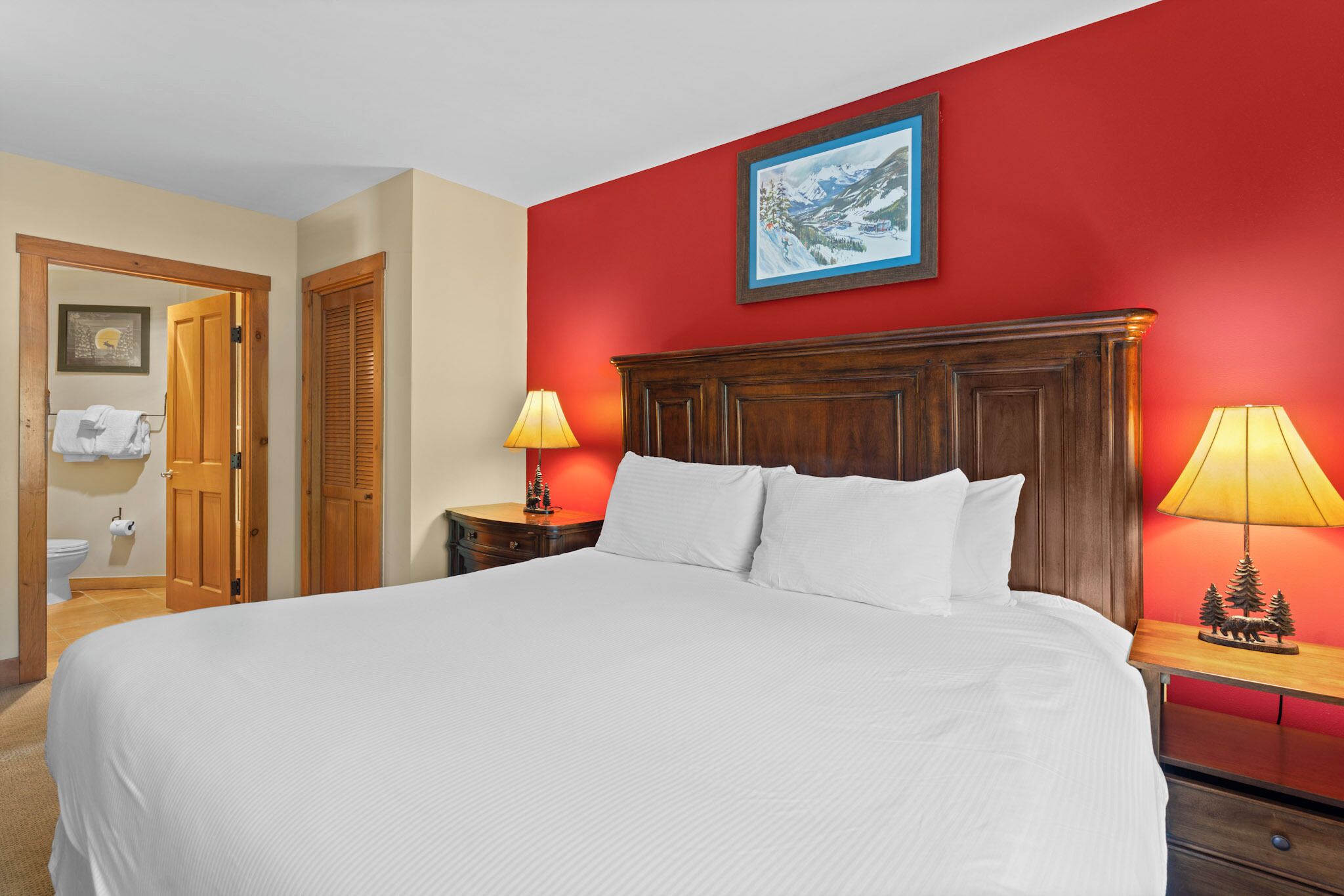 A hotel room with a large bed, wooden headboard, two bedside lamps, a painting on the wall, and a door leading to the bathroom.