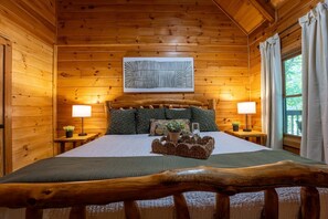A wooden cabin bedroom with a bed, two bedside lamps, a wall art piece, and a window with curtains.