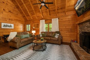 A cozy log cabin interior with a wooden ceiling, a fireplace, a sofa set, and a ceiling fan.