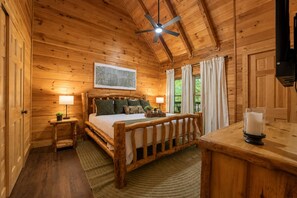 A wooden cabin bedroom with a bed, bedside lamps, a ceiling fan, and a window with curtains. - Cozy 1BR cabin in Pigeon Forge with hot tub, fireplace, arcade, pool access. (Pigeon Forge)
