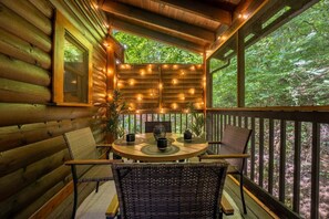 A wooden log cabin with a round table and chairs, string lights, and a view of the forest.