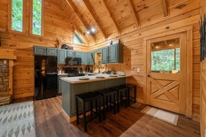 A wooden cabin interior with a kitchen area featuring a central island, bar stools, and a stone accent wall. - Cozy 1BR cabin in Pigeon Forge with hot tub, fireplace, arcade, pool access. (Pigeon Forge)