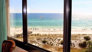 A room with a large window overlooking a beach with a lifeguard tower and a parasail in the distance.