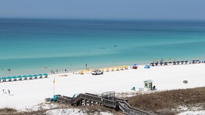 A beach with white sand, clear turquoise water, and a wooden boardwalk leading to beach umbrellas and a lifeguard tower.