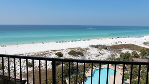 A beach view from a balcony with a pool, beach umbrellas, and a lifeguard tower.