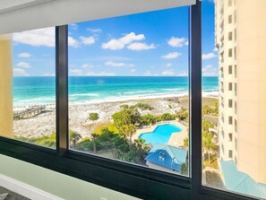 A view from a window overlooking a beach with a clear sky, a swimming pool, and a building in the background.