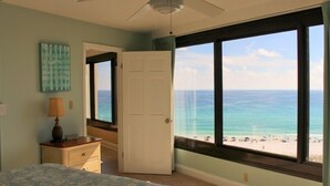 A bedroom with a bed, a nightstand, a lamp, and a view of the ocean through large windows.