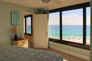 A bedroom with a bed, a nightstand, a lamp, and a view of the ocean through large windows.