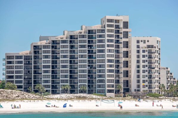 A multi-story hotel with balconies, located near a beach with people and umbrellas.