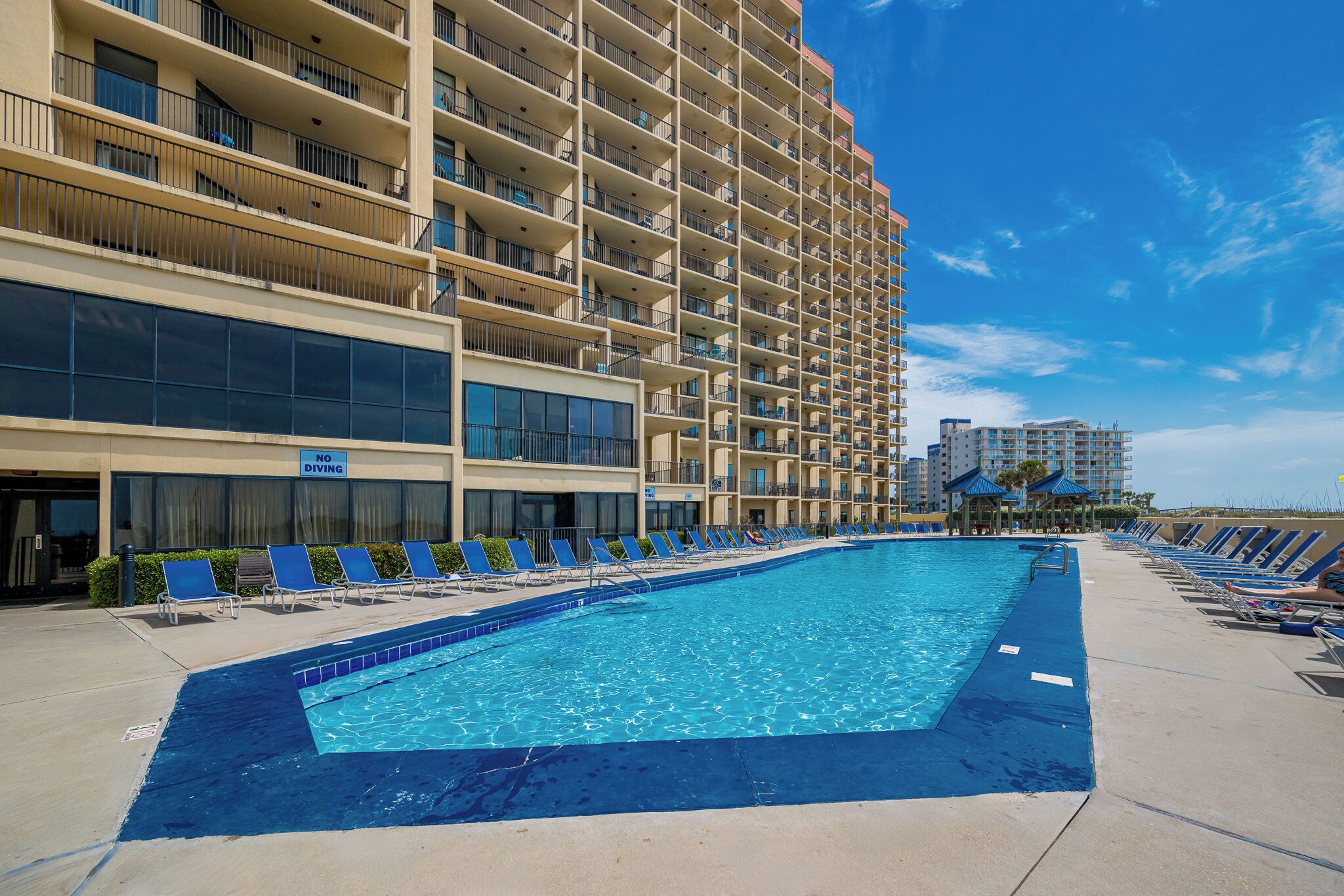 A hotel with a large swimming pool, multiple balconies, and a clear blue sky.