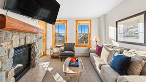 A living room with a stone fireplace, a flat-screen TV mounted above it, a sofa, a coffee table with a vase, and two windows with wooden frames. - 3 min Walk to Gondola, Private Balcony w/ Decrum Square & Mountain Views (Keystone)