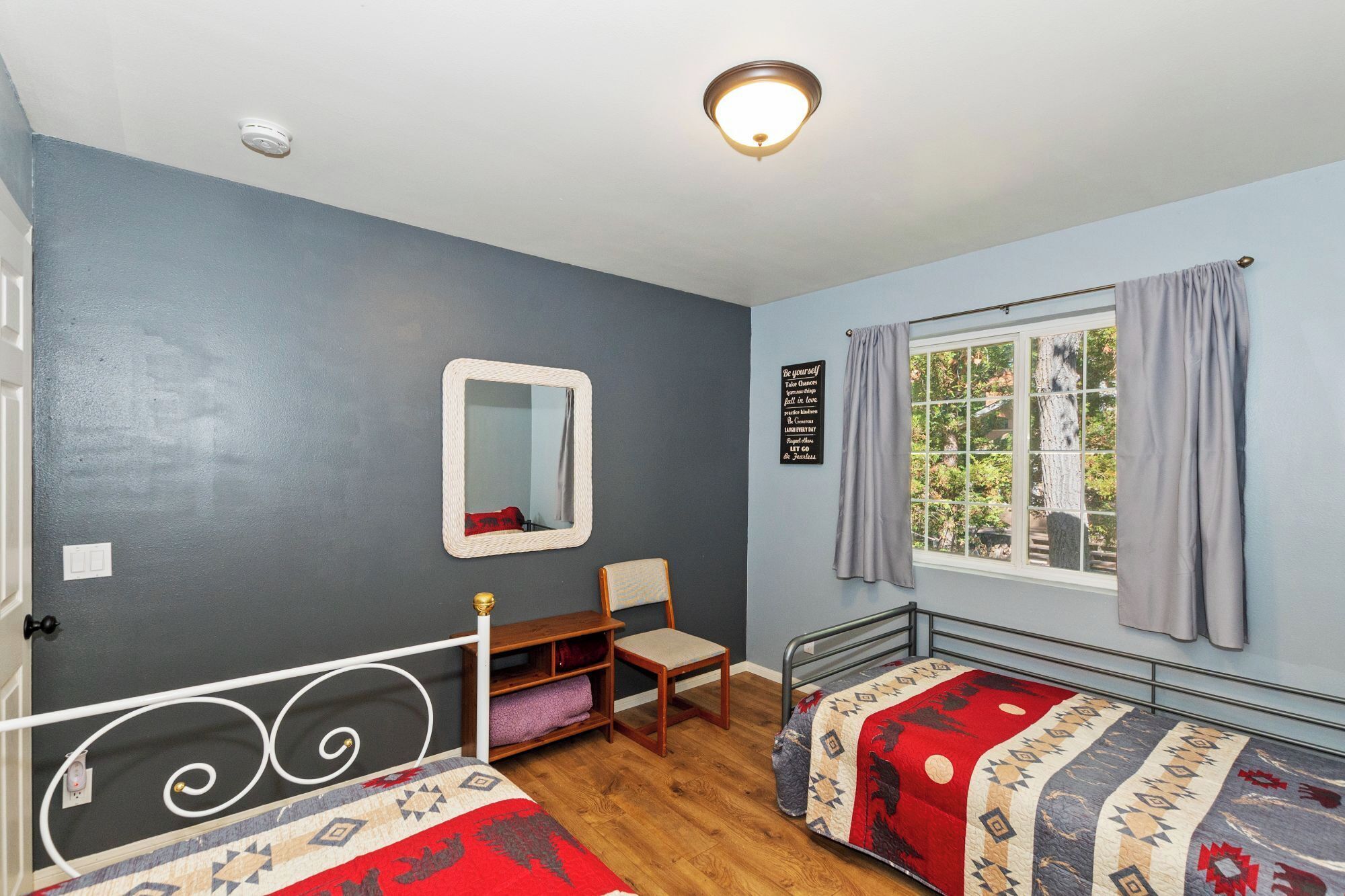 A bedroom with two beds, a wooden chair, a small table, a mirror, a window with curtains, and a motivational quote on the wall.
