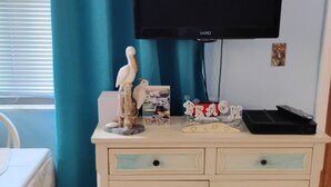 A room with a white cabinet featuring a beach scene, a flat-screen TV mounted on the wall, and a small table with a white tablecloth. -  Nice Condo directly on St Pete beach, walk to  restaurants/night life.  (St Pete Beach)
