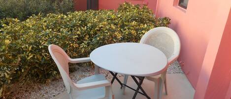 Un área de estar al aire libre con una mesa blanca y dos sillas blancas, rodeada por un seto y un muro de edificio rosa.