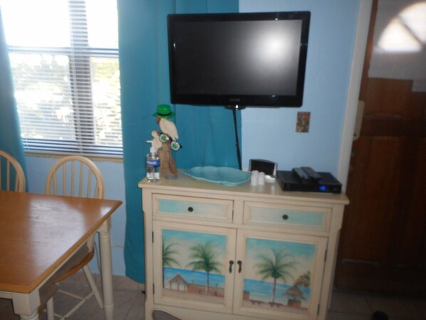 Una habitación con una televisión montada en la pared, una mesa de madera, sillas, un armario con puertas decorativas y una ventana con persianas.