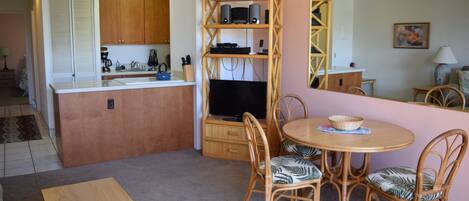 A compact kitchen with wooden cabinets, a built-in TV, and a dining area with wicker chairs and a table.