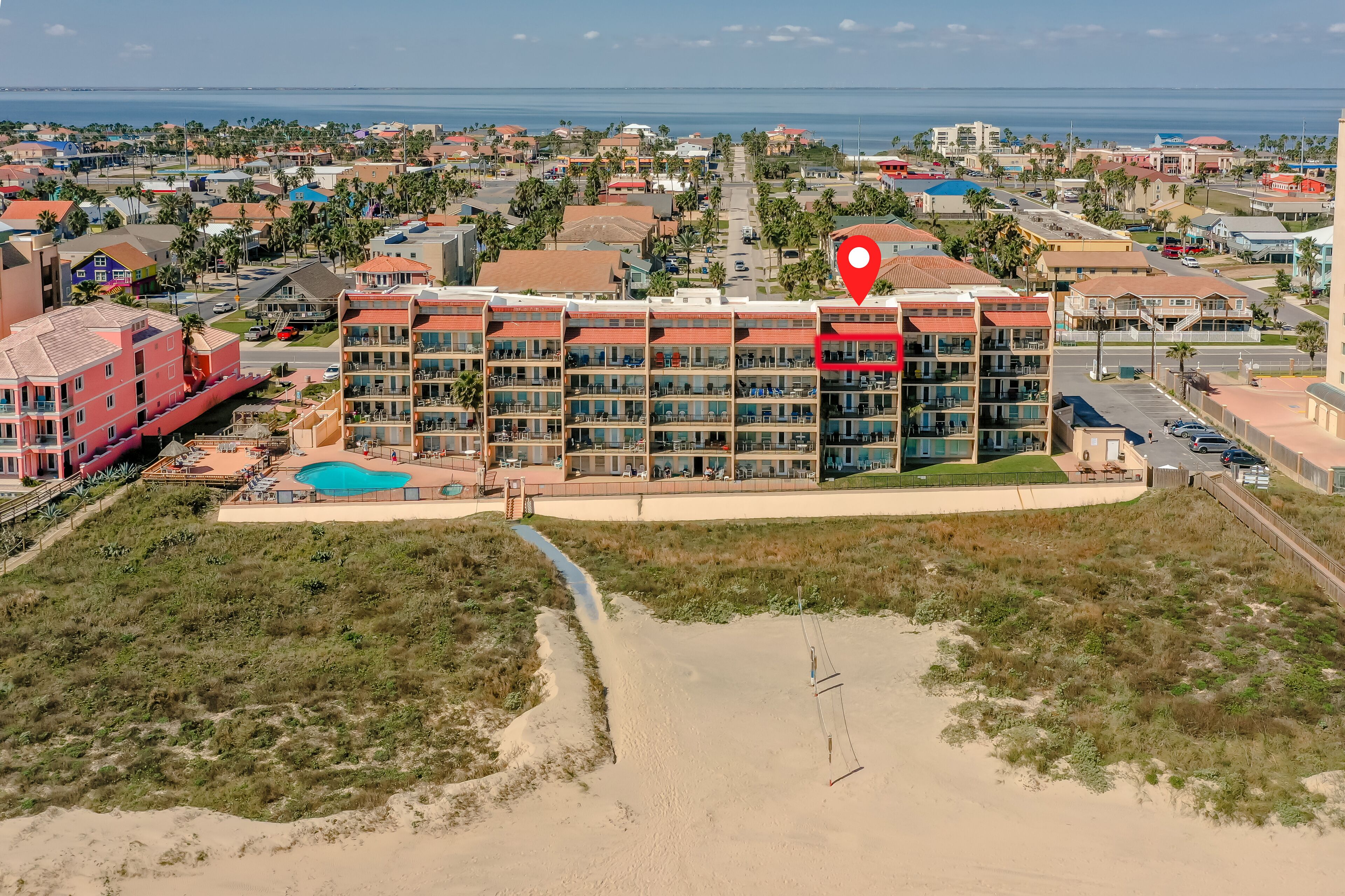 Een meerlaags hotel met zwembad en toegang tot het strand, omringd door woongebouwen en een strand.