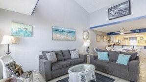 Een woonkamer met twee grijze banken, een witte rieten salontafel en een gestreept vloerkleed. Er hangen kunstwerken aan de muur en er is een plafondventilator.