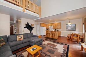A spacious living area with a gray sectional sofa, a wooden coffee table, and a patterned rug. A kitchen with wooden cabinets and a dining area with a wooden bench and table are visible.