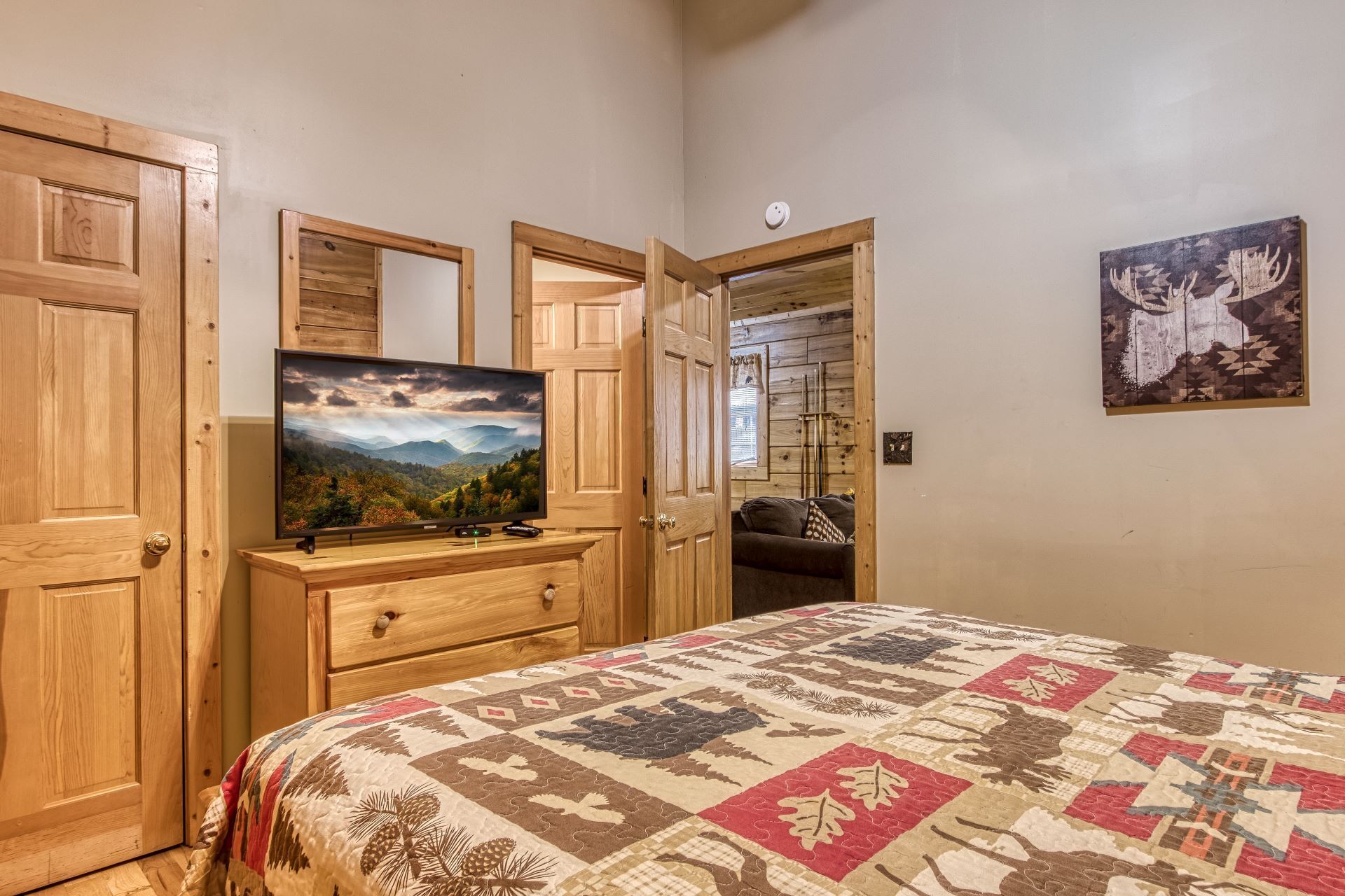A bedroom with a wooden dresser, a television on top, a quilted bedspread, and a painting on the wall.