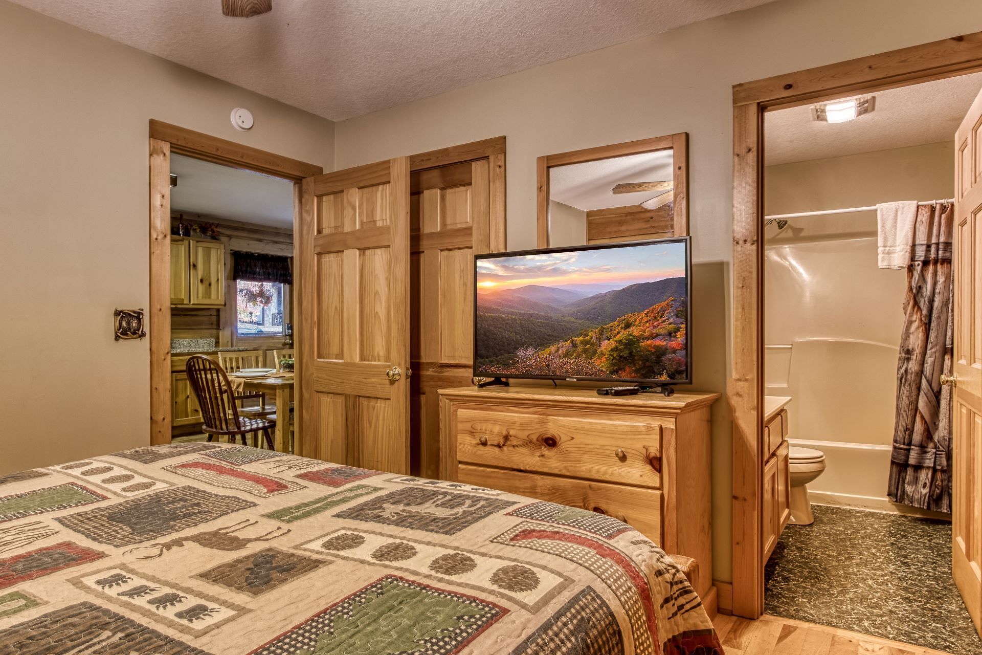 A bedroom with a bed, a television, a wooden dresser, a bathroom with a toilet and shower, and a window with a view of a mountainous landscape.