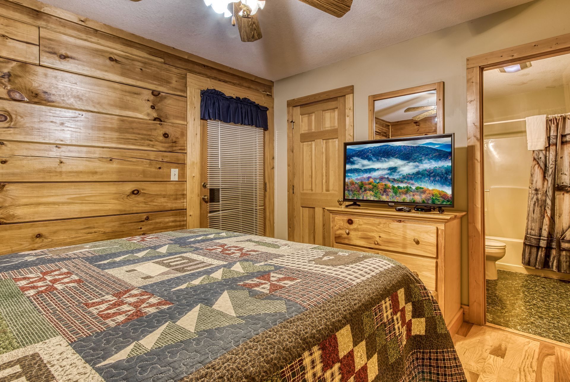 Una habitación acogedora con paredes de madera, una cama con colcha, un televisor sobre un aparador de madera y un baño visible a través de una puerta abierta.