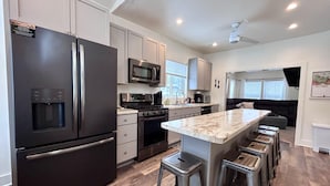 A modern kitchen with stainless steel appliances, a central island with bar stools, and a view to the living area.