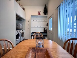 Un coin repas avec une table et des chaises en bois, un lave-linge encastré, une étagère avec des crochets et une fenêtre avec des stores.