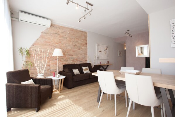 Een moderne woonkamer met een bakstenen muur, een donkerbruine bank, witte stoelen, een houten eettafel en een klein witte bijzettafeltje met een lamp.