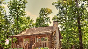 A rustic log cabin with a stone chimney, wooden porch, and a view of the surrounding forest. - ER65 - Eagles Point - Great location. Close to all the action! (Pigeon Forge)