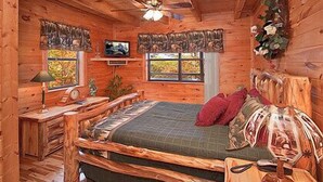 Un dormitorio de cabaña de madera con una cama, un televisor, un ventilador de techo y una ventana con vistas a árboles.