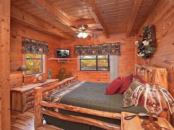 Une chambre dans une cabane en bois, équipée d’un lit, d’une télévision, d’un ventilateur de plafond et d’une fenêtre donnant sur la forêt.