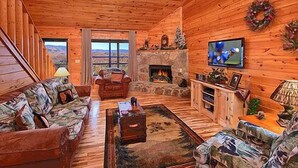 Interior de una cabaña de troncos con chimenea, televisor y vistas a las montañas a través del ventanal.