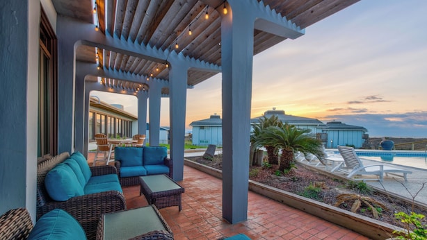 A covered outdoor seating area with wicker furniture and a view of a pool and buildings in the background. - Amazing sunrise views, swimming pool, boardwalk to the beach, picnic tables, pla (Port Aransas)