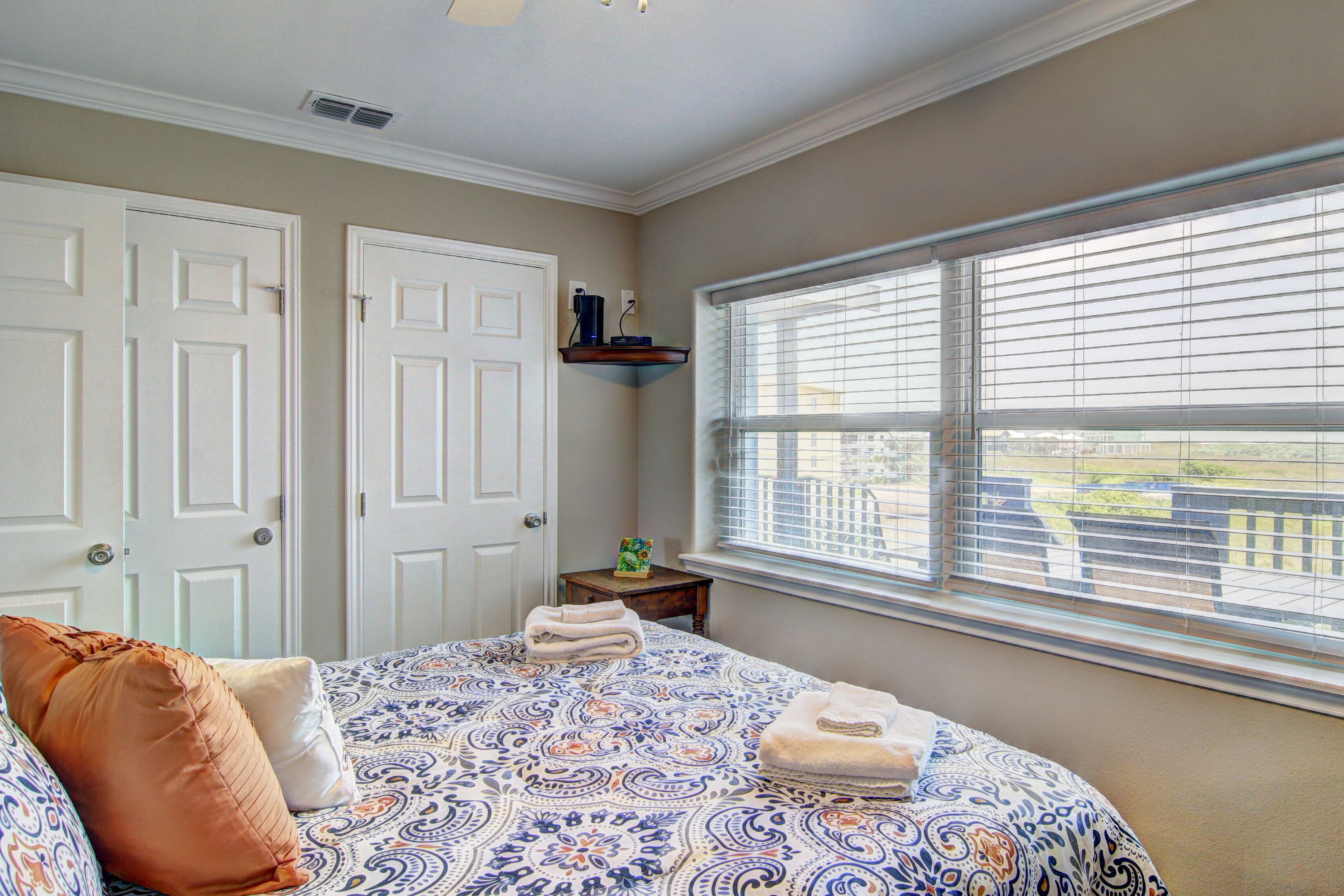 A bedroom with a patterned bedspread, two doors, a window with blinds, and a small table with a decorative item.