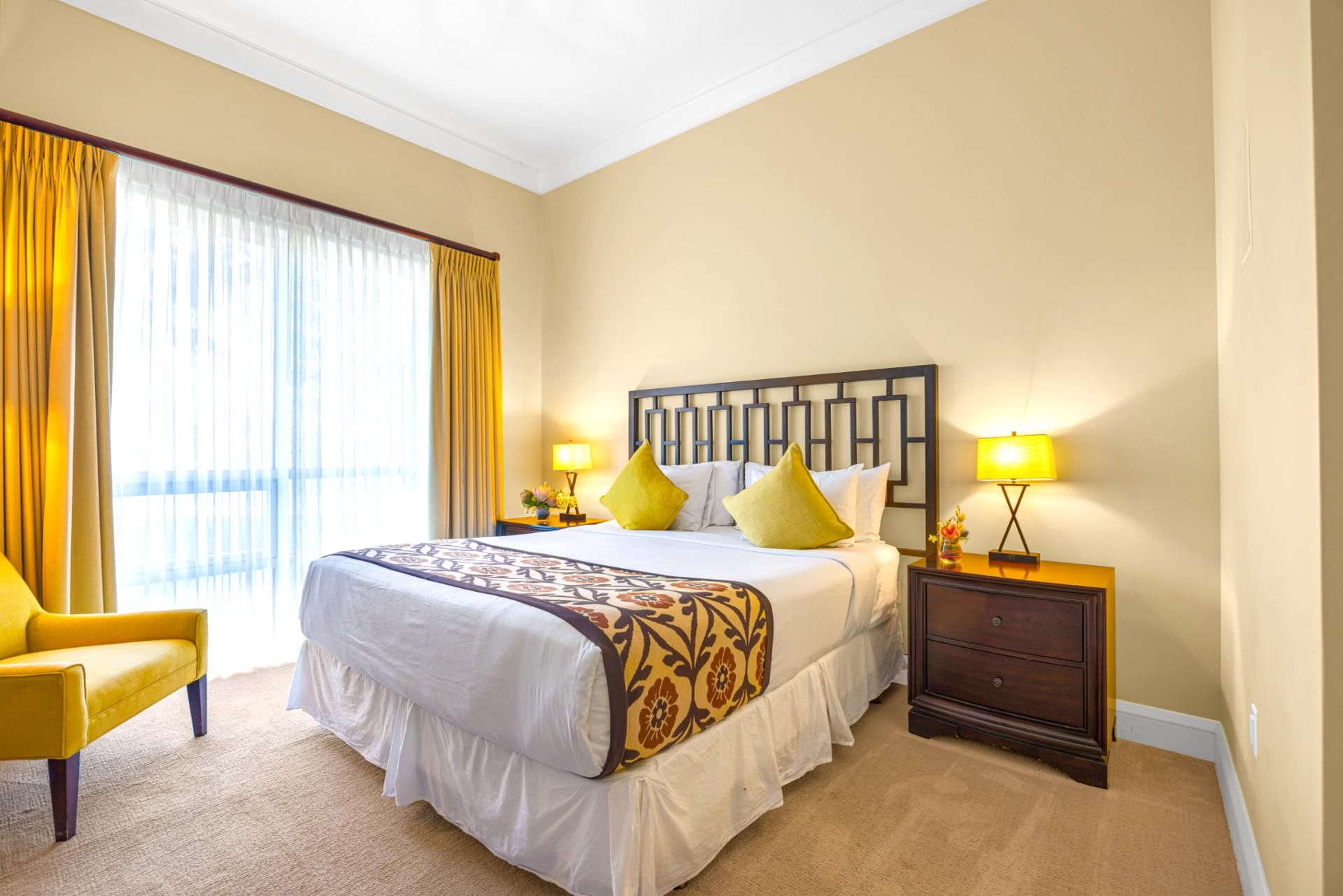A hotel room with a large bed, two bedside tables with lamps, a chair, and a view of the outdoors through large windows.