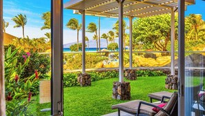 Ein überdachter Außenbereich mit Pergola, Liegestühlen und Blick auf eine tropische Landschaft mit Palmen und Strand.