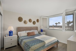 Un dormitorio con una cama, dos mesitas de noche, una ventana con contraventanas y vistas a palmeras.