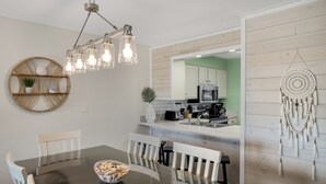 A modern kitchen with a central island, a dining table with chairs, and a decorative wall hanging. - 🌴 Islander #107 “Good Vibrations” – Your Easy, Fun Beach Getaway! 🌊 (Destin)