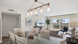 A modern living room with a dining area, a large window with a view, and a ceiling light fixture. - 🌴 Islander #107 “Good Vibrations” – Your Easy, Fun Beach Getaway! 🌊 (Destin)