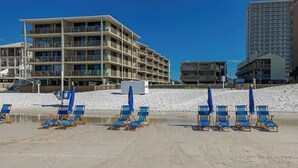 Un hotel sulla spiaggia con numerosi balconi, una spiaggia sabbiosa con sedie a sdraio blu e un cielo azzurro limpido.