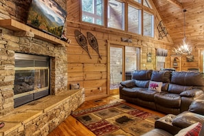 Cozy cabin-style room with a stone fireplace, leather sofa, and large window offering a view of the surrounding landscape.