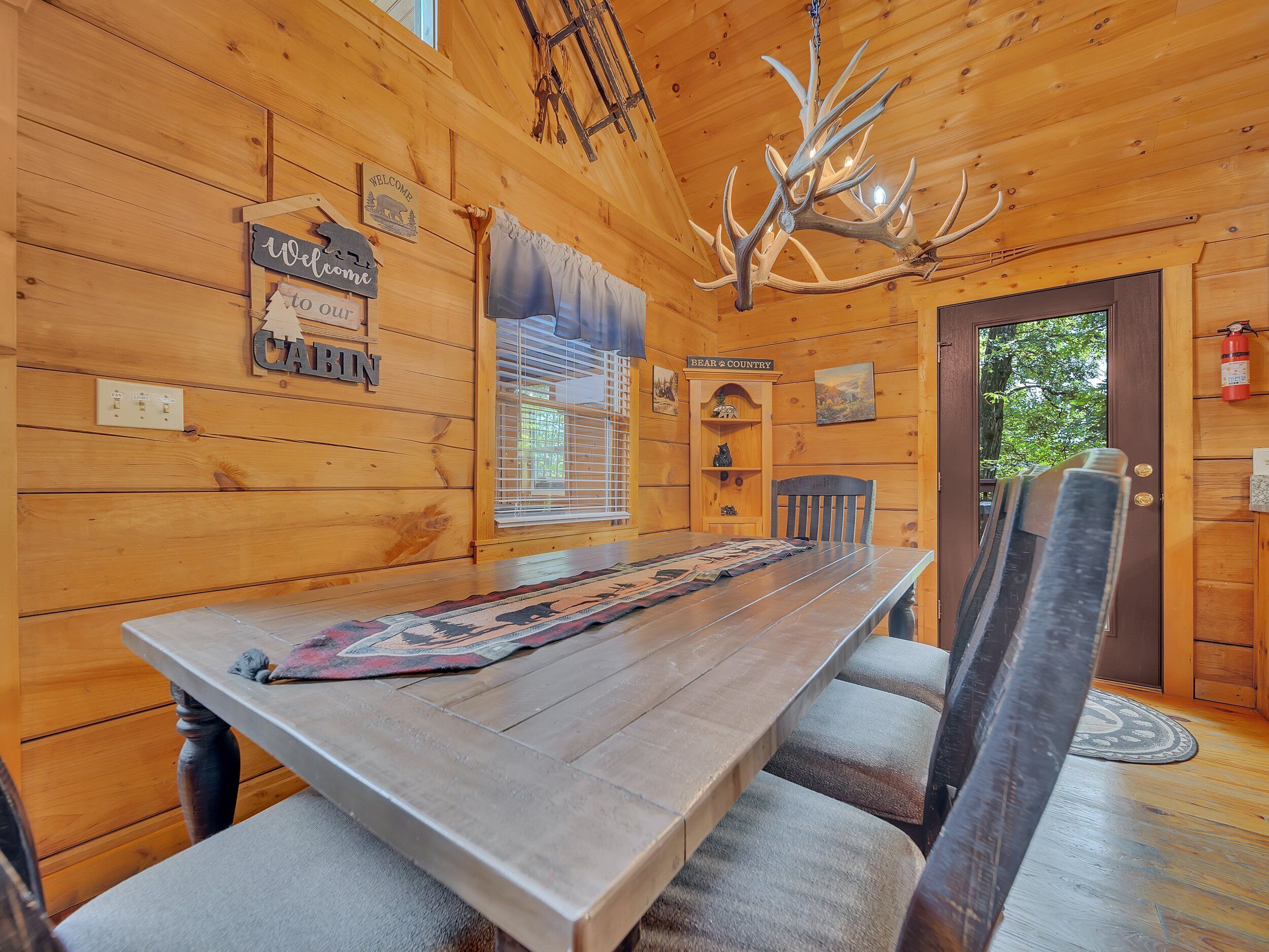 A wooden cabin interior with a long dining table, benches, a chandelier with antlers, and a 'Welcome to Cabin' sign.