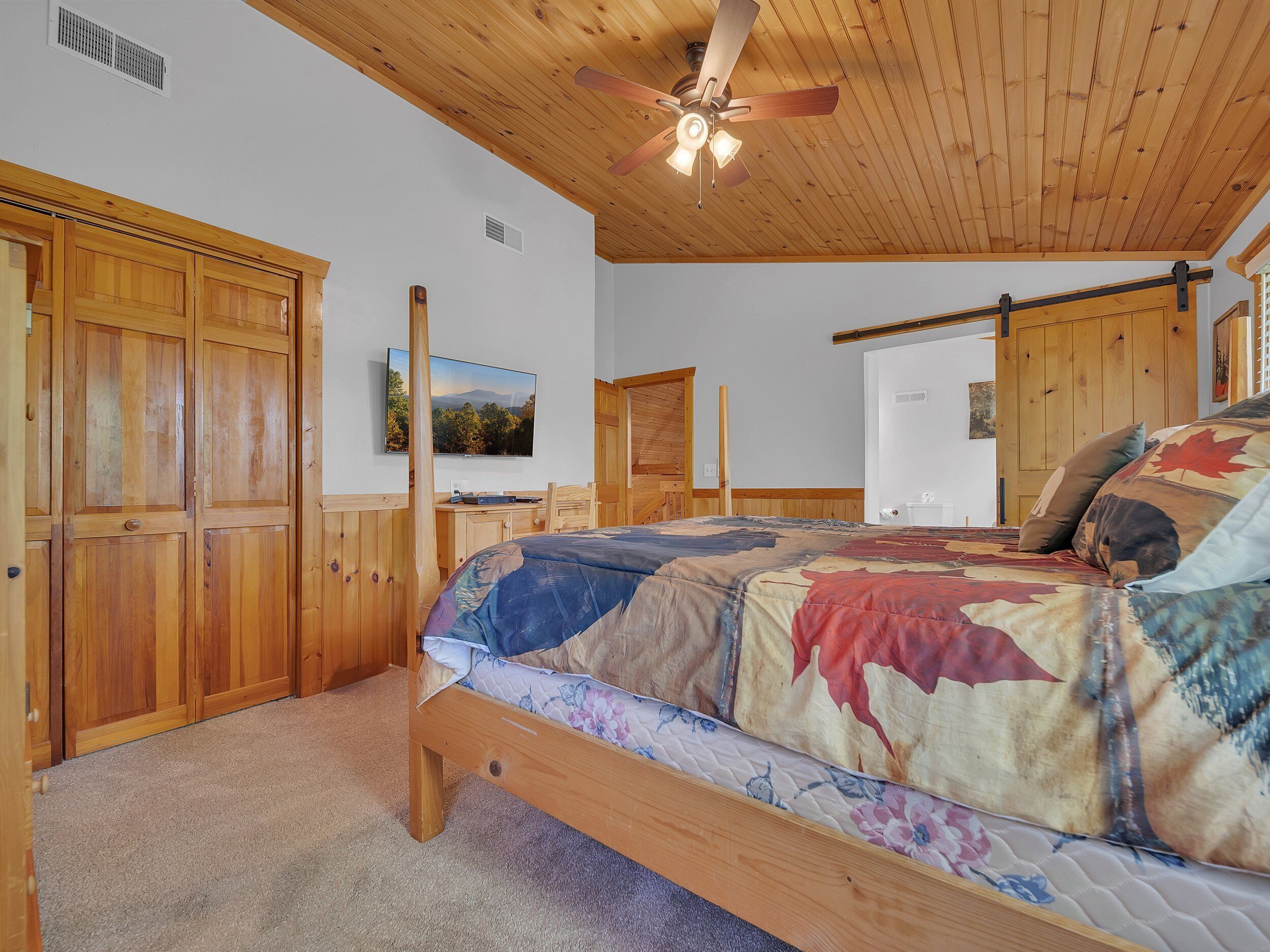 A bedroom with a wooden ceiling, a bed with a floral comforter, a television on the wall, and wooden cabinets.