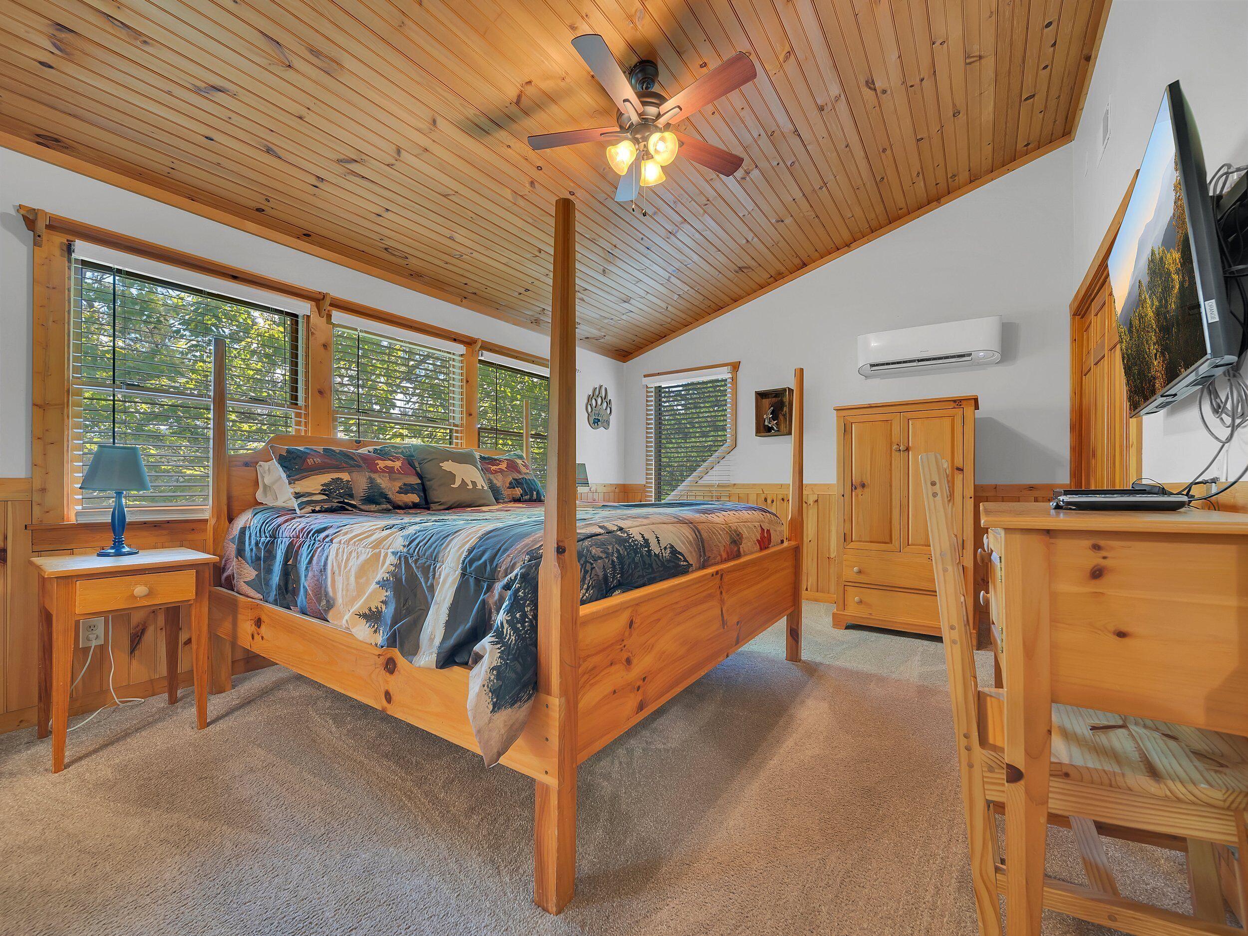 A wooden bedroom with a bed, a desk, a ceiling fan, and large windows.