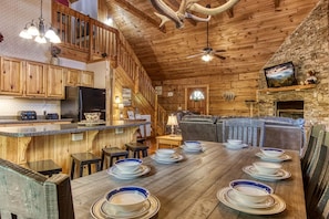 Cozy cabin interior with a warm aesthetic, featuring a kitchen with wooden cabinets, a dining table set for a meal, and a comfortable living area with a leather sofa and fireplace.