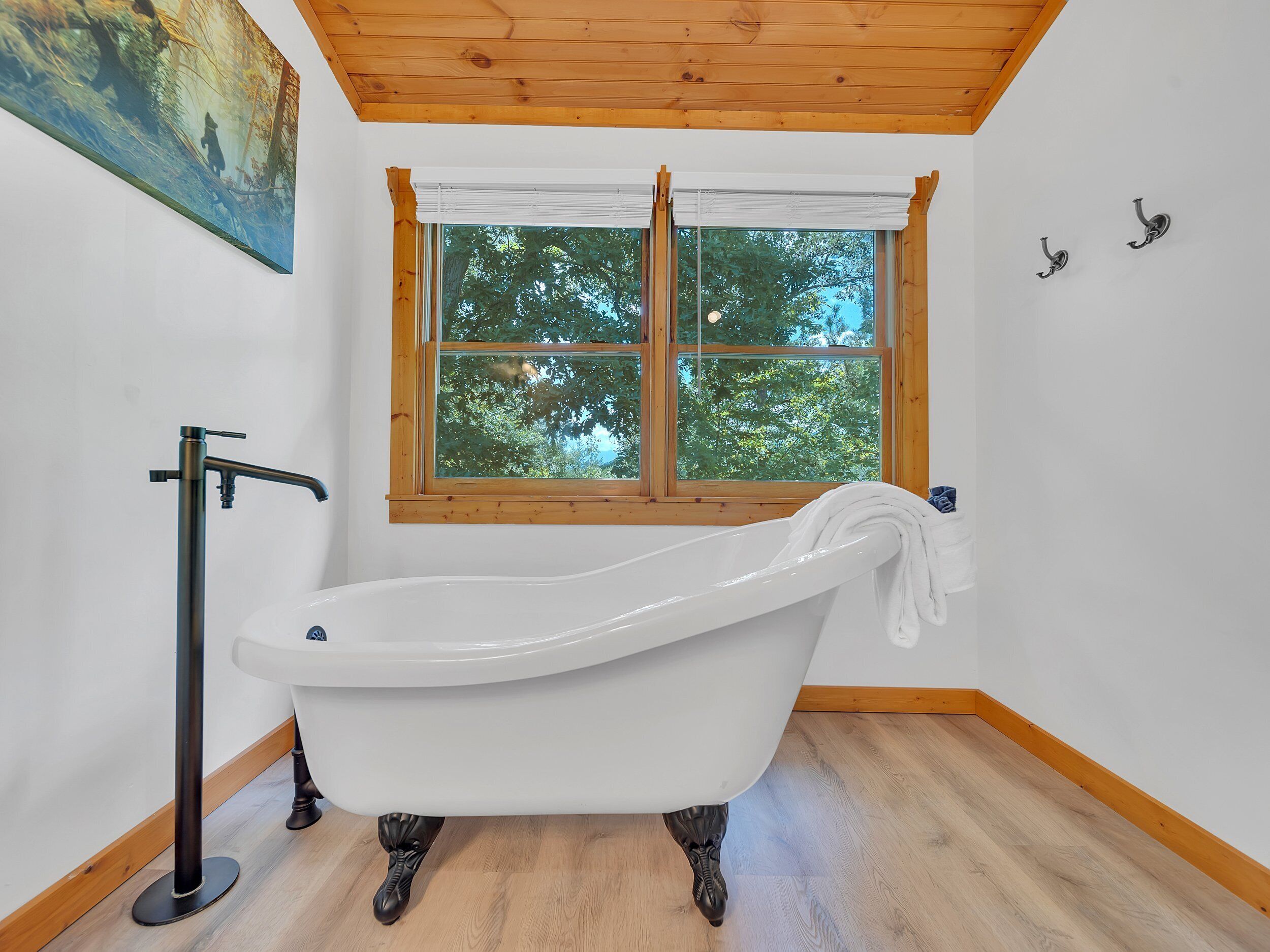 Un baño con bañera tipo pata de león, suelo de madera y ventana con vistas a árboles.