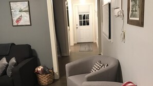 Un salon avec un canapé gris, un fauteuil gris et un plaid à carreaux rouges et blancs. Un tableau encadré est accroché au mur, et une porte donne accès à une autre pièce.